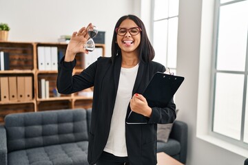 Canvas Print - Young hispanic woman working at consultation office holding sand clock sticking tongue out happy with funny expression.