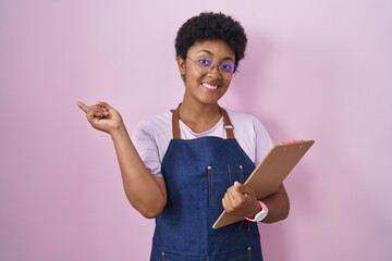 Poster - Young african american woman wearing professional waitress apron holding clipboard with a big smile on face, pointing with hand finger to the side looking at the camera.