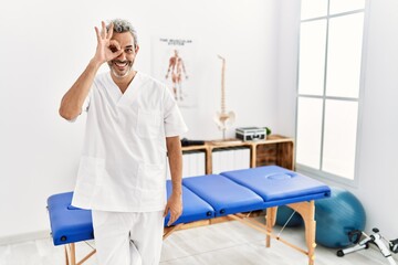 Sticker - Middle age hispanic therapist man working at pain recovery clinic doing ok gesture with hand smiling, eye looking through fingers with happy face.