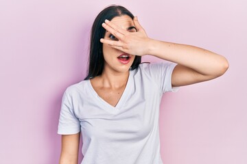 Poster - Beautiful woman with blue eyes wearing casual white t shirt peeking in shock covering face and eyes with hand, looking through fingers afraid