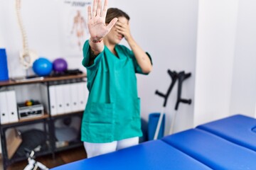 Poster - Young hispanic woman wearing physiotherapist uniform standing at clinic covering eyes with hands and doing stop gesture with sad and fear expression. embarrassed and negative concept.