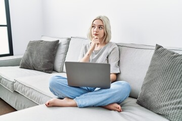 Poster - Young caucasian woman using laptop at home sitting on the sofa looking confident at the camera with smile with crossed arms and hand raised on chin. thinking positive.