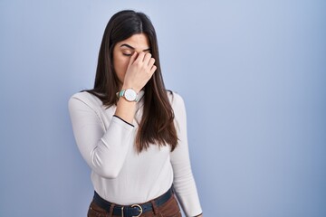 Canvas Print - Young brunette woman standing over blue background tired rubbing nose and eyes feeling fatigue and headache. stress and frustration concept.