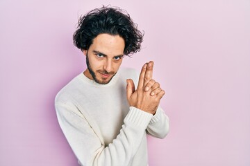 Sticker - Handsome hispanic man wearing casual white sweater holding symbolic gun with hand gesture, playing killing shooting weapons, angry face