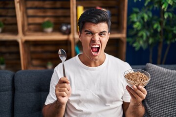 Canvas Print - Hispanic man eating healthy whole grain cereals with spoon angry and mad screaming frustrated and furious, shouting with anger. rage and aggressive concept.