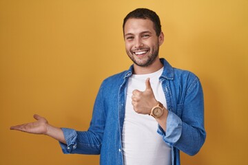 Hispanic man standing over yellow background showing palm hand and doing ok gesture with thumbs up, smiling happy and cheerful