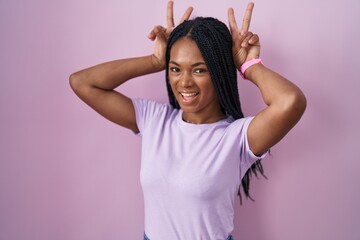 Poster - African american woman with braids standing over pink background posing funny and crazy with fingers on head as bunny ears, smiling cheerful