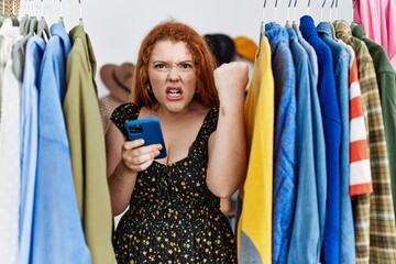 Poster - Young redhead woman searching clothes on clothing rack using smartphone annoyed and frustrated shouting with anger, yelling crazy with anger and hand raised