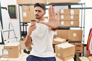 Wall Mural - Young hispanic man working at small business ecommerce drinking a coffee with open hand doing stop sign with serious and confident expression, defense gesture