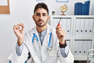 Poster - Young doctor man holding electronic cigarette at medical clinic relaxed with serious expression on face. simple and natural looking at the camera.