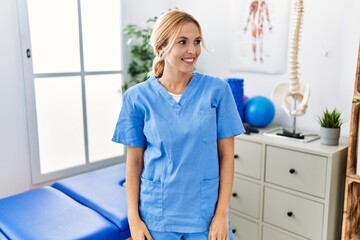 Poster - Beautiful blonde physiotherapist woman working at pain recovery clinic looking away to side with smile on face, natural expression. laughing confident.