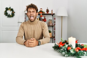 Poster - Young handsome man with beard sitting on the table by christmas decoration sticking tongue out happy with funny expression. emotion concept.