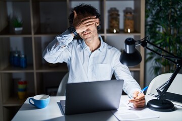 Canvas Print - Hispanic man working at the office at night smiling and laughing with hand on face covering eyes for surprise. blind concept.