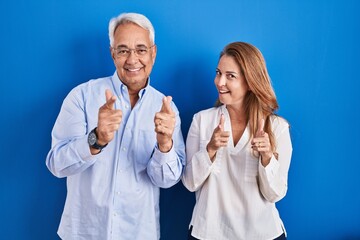 Poster - Middle age hispanic couple standing over blue background pointing fingers to camera with happy and funny face. good energy and vibes.