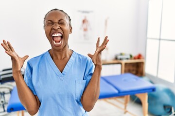 Wall Mural - Black woman with braids working at pain recovery clinic celebrating mad and crazy for success with arms raised and closed eyes screaming excited. winner concept