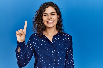 Poster - Young brunette woman with curly hair wearing casual clothes over blue background showing and pointing up with finger number one while smiling confident and happy.