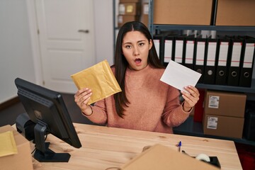 Canvas Print - Young latin woman working at small business ecommerce packing order in shock face, looking skeptical and sarcastic, surprised with open mouth