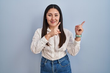 Sticker - Young latin woman standing over blue background smiling and looking at the camera pointing with two hands and fingers to the side.