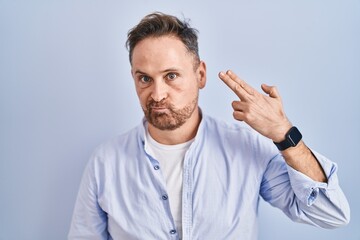 Sticker - Middle age caucasian man standing over blue background shooting and killing oneself pointing hand and fingers to head like gun, suicide gesture.