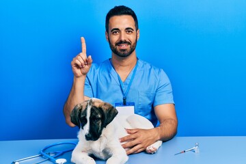 Wall Mural - Handsome hispanic veterinary man with beard checking dog health with a big smile on face, pointing with hand finger to the side looking at the camera.