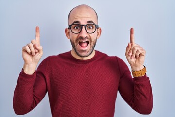 Sticker - Young bald man with beard standing over white background wearing glasses smiling amazed and surprised and pointing up with fingers and raised arms.