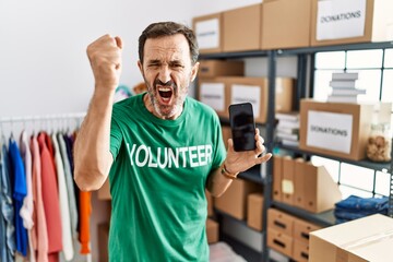 Canvas Print - Middle age man with beard wearing volunteer t shirt holding smartphone angry and mad raising fist frustrated and furious while shouting with anger. rage and aggressive concept.