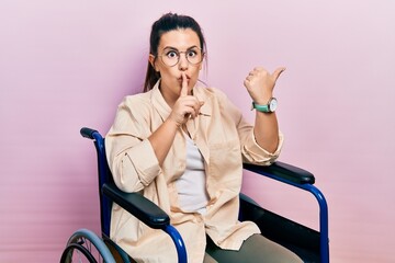 Sticker - Young hispanic woman sitting on wheelchair asking to be quiet with finger on lips pointing with hand to the side. silence and secret concept.