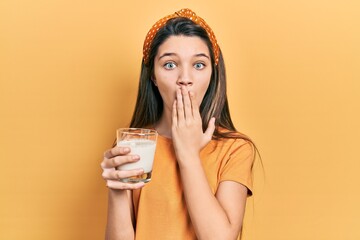 Wall Mural - Young brunette girl drinking a glass of milk covering mouth with hand, shocked and afraid for mistake. surprised expression