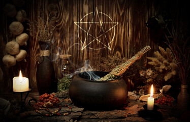 Witchcraft altar. Pentagram sign, burning candles, dry herbs, iron cauldron, smoking mugwort, bunch of garlic in wooden background in the dark, low key, selective focus.