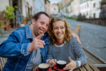 Portrait of happy young man with Down syndrome with his mother sitting at cafe outdoors and talking.