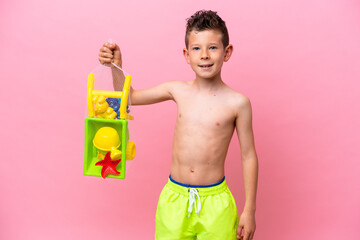 Wall Mural - Little caucasian boy holding beach toys isolated on pink background smiling a lot