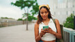 Cute tanned woman with long brown hair wearing white top and yellow bandana walks on bridge with backpack on her shoulder and cell phone in her hand. Girl using phone