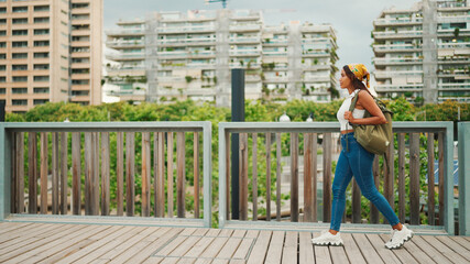 Wall Mural - Сute tanned woman with long brown hair wearing white top and yellow bandana walks on bridge with backpack on her shoulder and looks at modern city