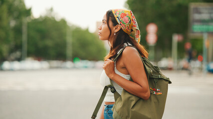 Wall Mural - Profile of cute tanned woman with long brown hair in white top and yellow bandana with backpack on her shoulders is walking along pedestrian crossing