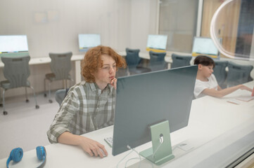 Wall Mural - A ginger schoolboy sitting at the computer at school and looking involved