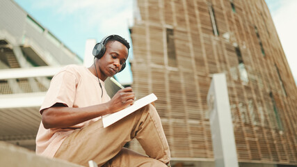 young african student sitting outside of university and taking notes while study online with headpho