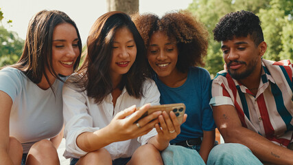 Wall Mural - Happy, smiling multiethnic young people at picnic on summer day outdoors. Group of friends talking, using cellphone while relaxing in the park at picnic