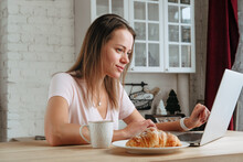 woman  working from home, eating a croissant and coffee for breakfast and looking at the laptop screen