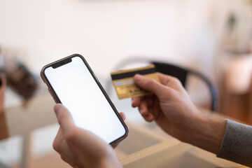 Close up young man holding bank credit card and modern tech device with mock up white screen in hands, enjoying purchasing goods in internet store using mobile phone application, online shopping
