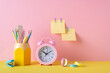 Back to school concept. Photo of colorful school supplies alarm clock adhesive tape pencil holder mini stapler and sticky note paper attached to pink wall