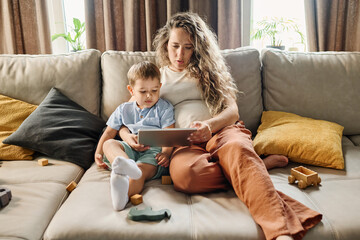 Young pregnant woman and little boy watching cartoons or movie for children or communicating in video chat while resting at home