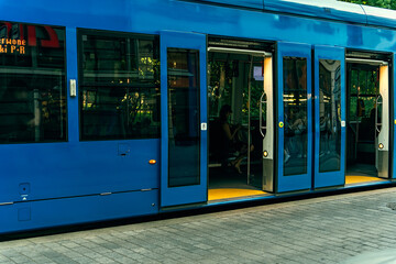 Blue tram with open doors for passengers at a stop on a summer evening, reflections in the windows of the tram, sunlight on the branches of trees outside the windows of the tram, sustainable transport