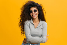 Female Portrait. Happy Latin Woman With Afro In Sunglasses With Folded Arms Smiling At Camera, Wearing T-shirt, Posing Isolated Over Yellow Studio Background. Positive Casual Female Model Laughing