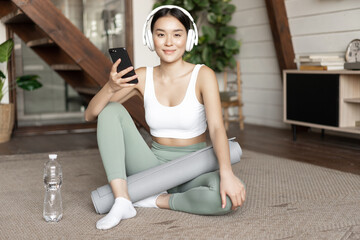 Wall Mural - Smiling asian girl in headphones wearing sport clothing, listening music, holding mobile phone and workout at home alone, sitting in living room