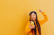 happy asian young woman in yellow sweater listening to music with a headphone and holding her smartphone with yellow wall background.