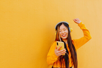 happy asian young woman in yellow sweater listening to music with a headphone and holding her smartp