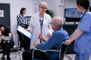 Wall Mural - Senior doctor talking with retired person using wheelchair about appointment while being helped by professional nurse in private clinic. Older man living with disability in busy hospital reception.