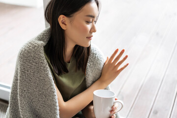 Wall Mural - Thoughtful asian woman sitting at home on floor, touching glass window nostalgic, yearning to go outside, being on self-quarantine