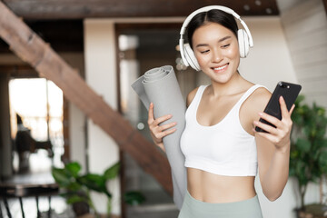 Smiling asian girl with headphones, looking at mobile phone and holding rubber mat for workout, fitness at home, concept of lifestyle and sport