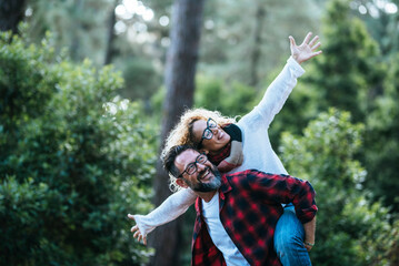 Overjoyed and happy couple have fun together in the park nature. Man carry woman in piggyback in outdoor leisure activity. Green forest in background. Hipster person smile and laugh a lot outdoors